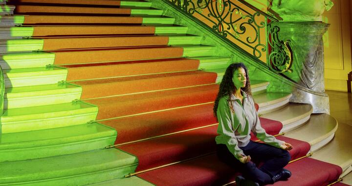 Eine Frau meditiert auf einer großen Treppe in einem historischen Gebäude mit roter und grüner Beleuchtung.