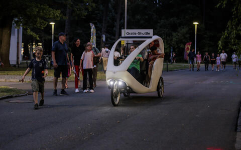 A pedal-powered shuttle labeled “Gratis-Shuttle” carries passengers at night.