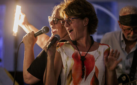 Two women passionately sing into microphones at a nighttime event.