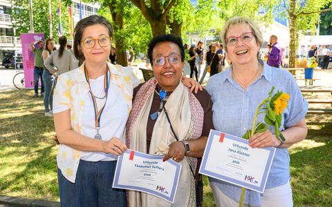 Three women holding award certificates and smiling at an outdoor celebration.