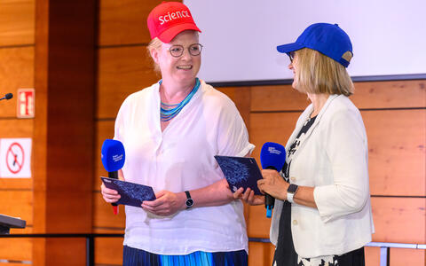 Two women wearing science-themed caps speak on stage with branded microphones.