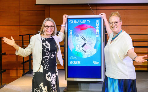 Two women pose next to a digital sign for Summer Science Day 2025 at Max Delbrück Center.