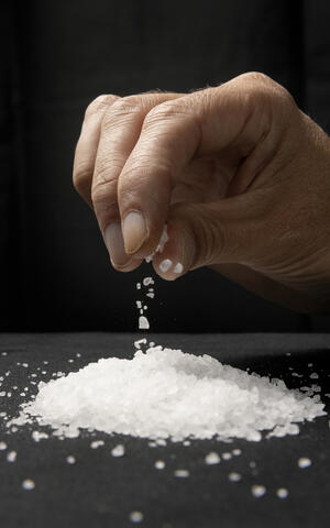 Eine Hand salzt über einem Salzhäufchen auf dem schwarzem Hintergrung
