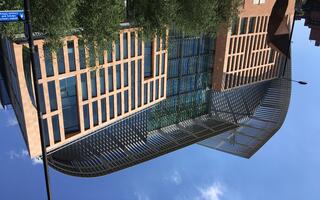 Modern building complex with glass façade and striking metal structure on the roof, surrounded by trees, under a blue sky.
