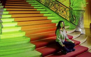 Eine Frau meditiert auf einer großen Treppe in einem historischen Gebäude mit roter und grüner Beleuchtung.