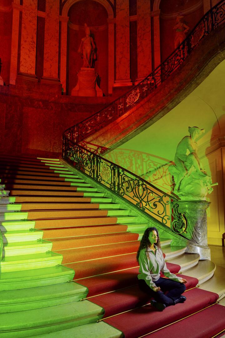Woman meditating on a grand staircase lit with red and green lighting in a historic building.