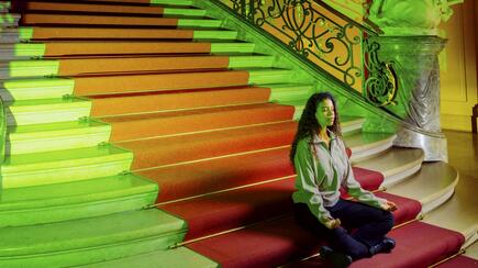 Woman meditating on a grand staircase lit with red and green lighting in a historic building.